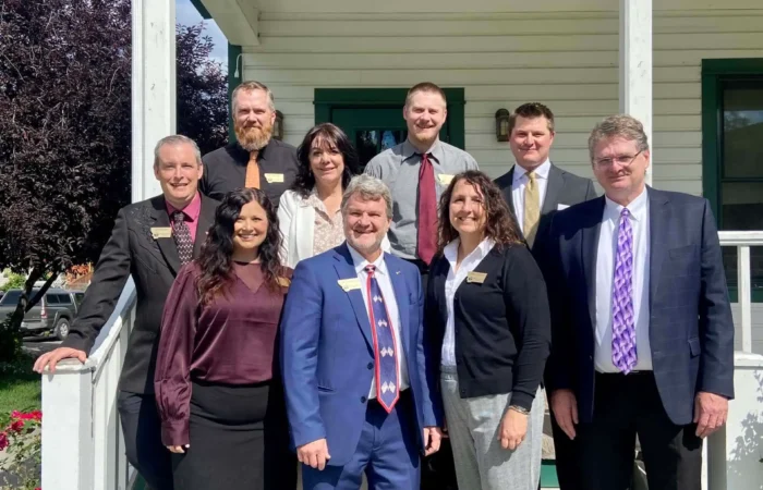 Secretary of State's Office presents at 2023 Montana Association of Clerks & Recorders Convention 6 A group of nine adults in business attire stand together and smile on the steps of a white house with green trim on a sunny day, gathered for the Montana Association Clerks & Recorders Convention. Some greenery and a tree are visible in the background.