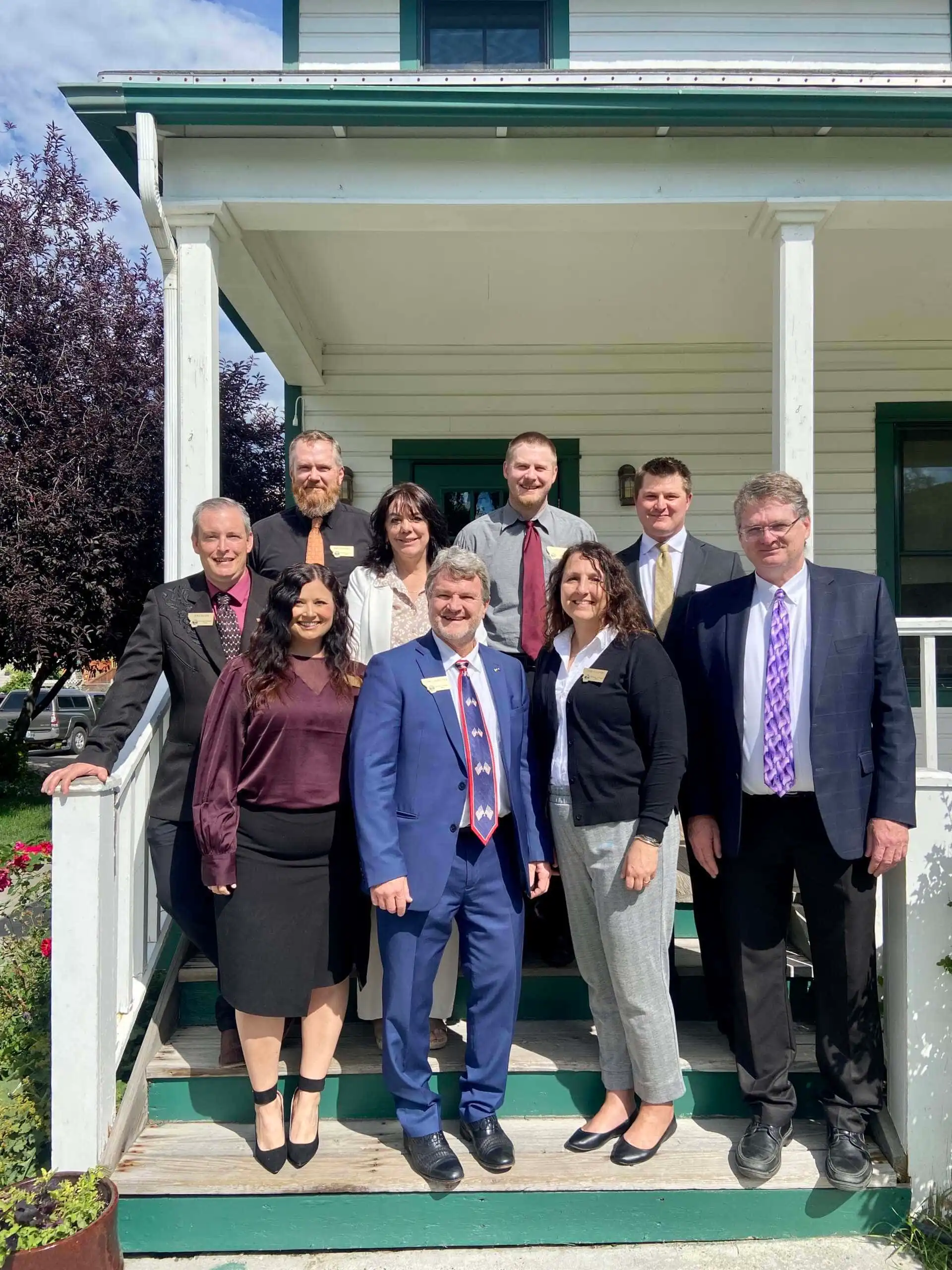 A group of nine adults in business attire stand together and smile on the steps of a white house with green trim on a sunny day, gathered for the Montana Association Clerks & Recorders Convention. Some greenery and a tree are visible in the background.