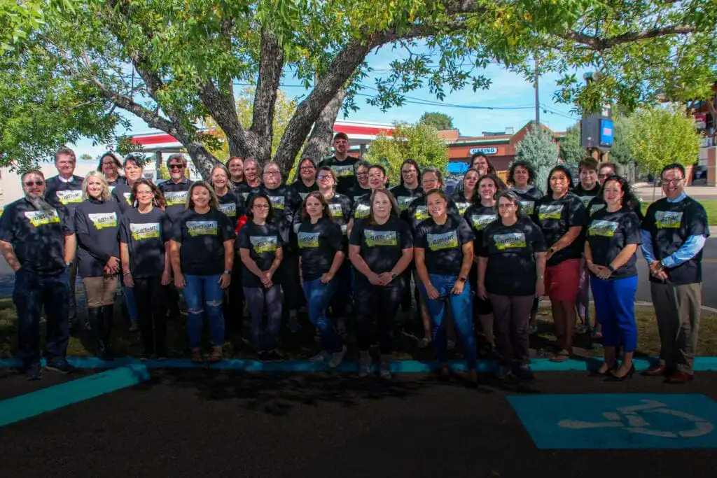 A large group of people wearing matching black shirts stands outdoors in front of a tree in Great Falls, smiling at the camera. There are buildings, street signs, and parked cars visible in the background during electMT training.