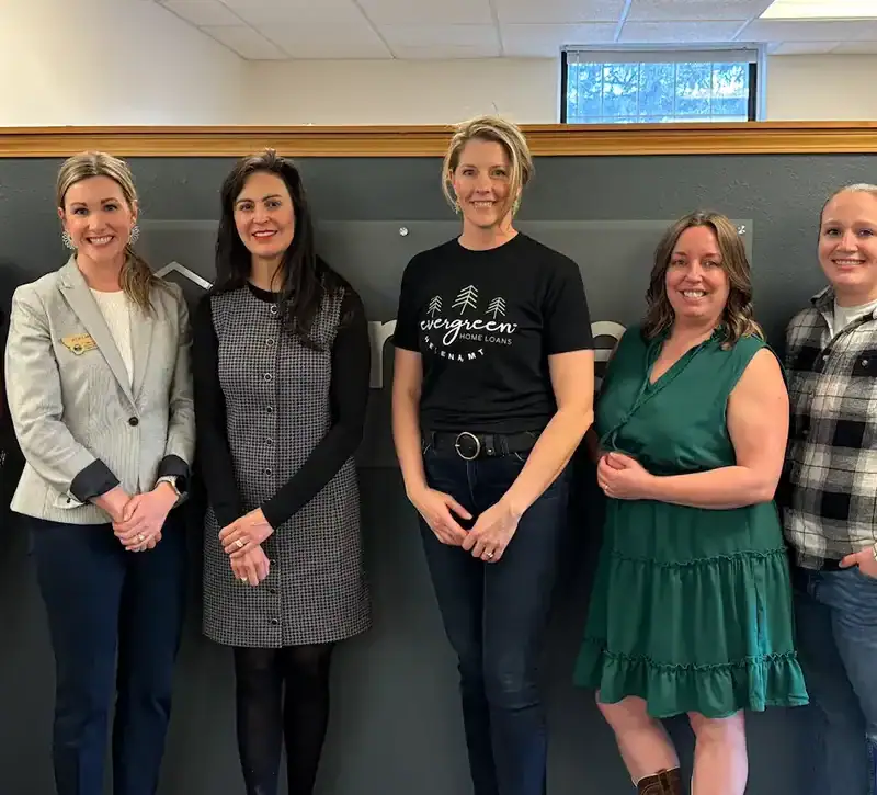 Seven people stand indoors in a row, smiling at the camera. The group includes men and women dressed in business casual attire. The office setting with gray walls and a window reflects the team’s focus on new business registrations in March 2025.