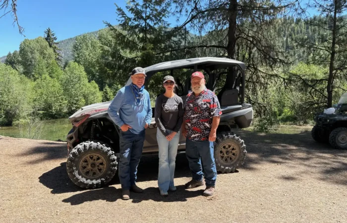 Secretary Christi Jacobsen grateful for tour of Fish Creek, shedding light on future of recreation in Mineral County 5 Three people stand smiling in front of an off-road vehicle on a sunny day near Fish Creek, enjoying recreation in Mineral County, surrounded by trees and greenery.