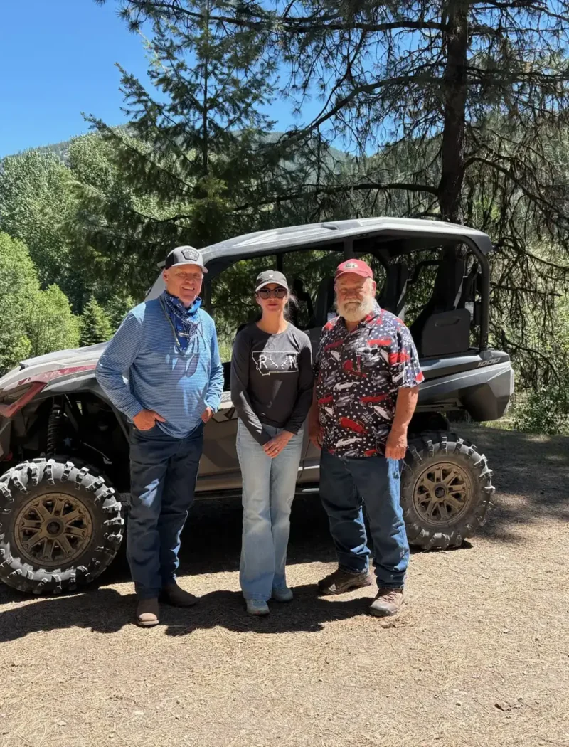 Three people stand smiling in front of an off-road vehicle on a sunny day near Fish Creek, enjoying recreation in Mineral County, surrounded by trees and greenery.