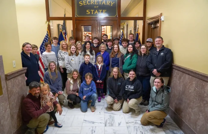 Secretary Christi Jacobsen meets with Foothills Community Christian School students, staff 5 A large group of adults and Foothills Community Christian School students pose for a photo inside a government building in front of a door labeled Secretary of State, with American flags on either side and marble floors beneath them.
