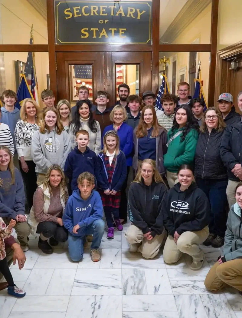 Secretary Christi Jacobsen meets with Foothills Community Christian School students, staff 1 A large group of adults and Foothills Community Christian School students pose for a photo inside a government building in front of a door labeled Secretary of State, with American flags on either side and marble floors beneath them.