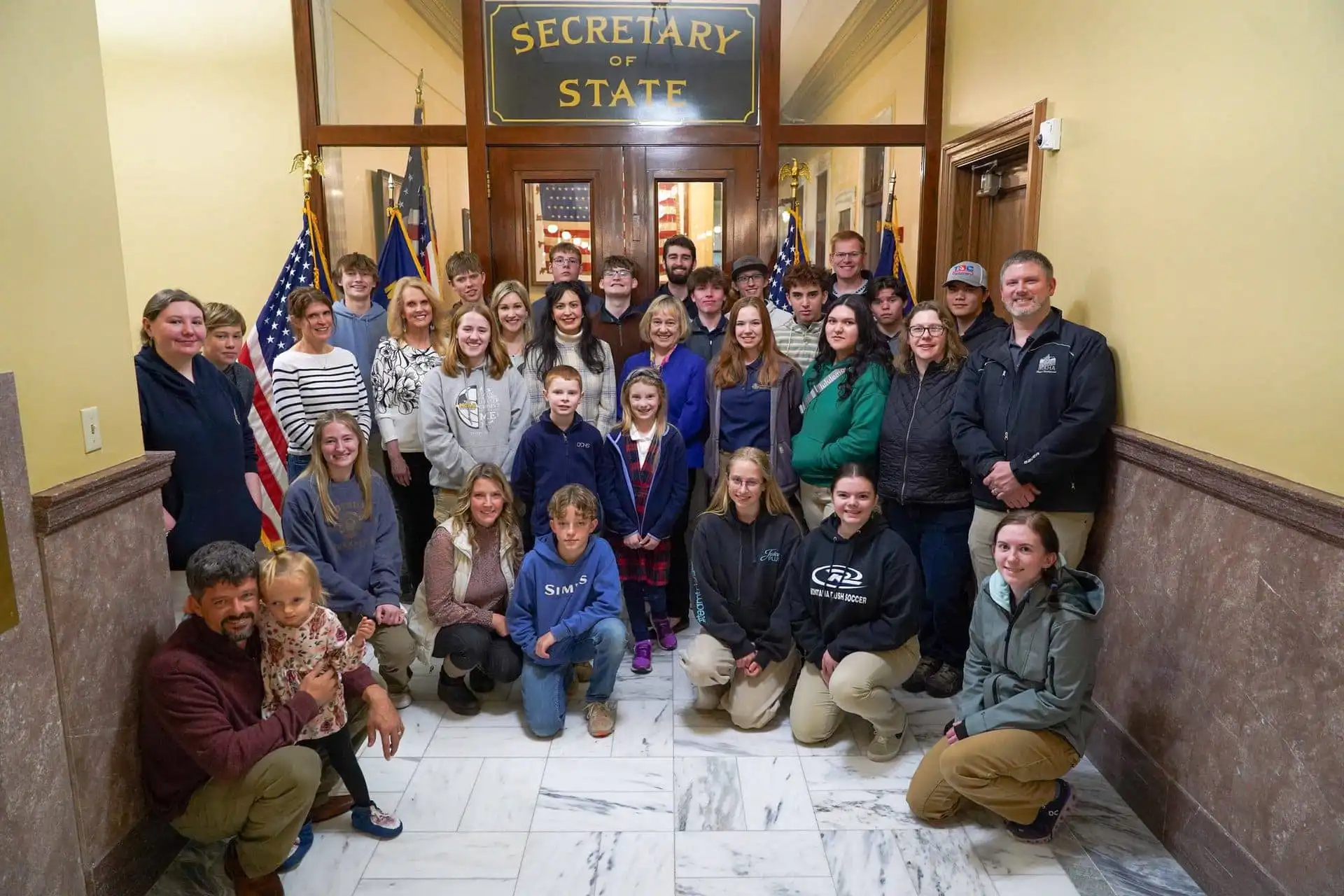 Secretary Christi Jacobsen meets with Foothills Community Christian School students, staff 10 A large group of adults and Foothills Community Christian School students pose for a photo inside a government building in front of a door labeled Secretary of State, with American flags on either side and marble floors beneath them.
