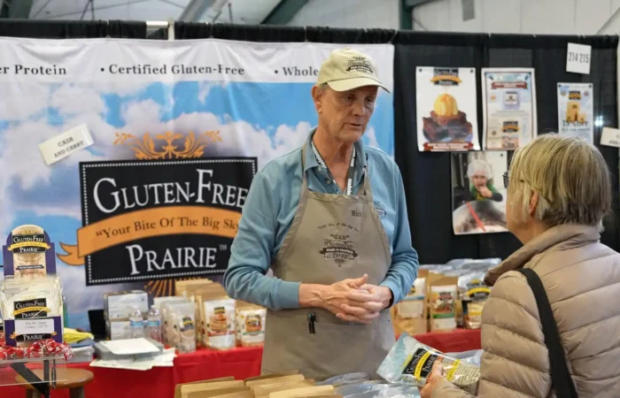 A vendor in a beige apron and cap talks to a customer at a booth labeled Gluten-Free Prairie during the Made in Montana Tradeshow. Various gluten-free products from Montana businesses are displayed, with banners and packages visible in the background.