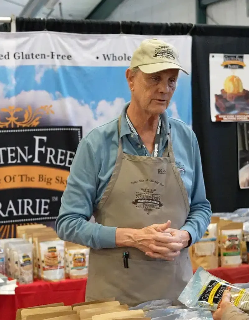 A vendor in a beige apron and cap talks to a customer at a booth labeled Gluten-Free Prairie during the Made in Montana Tradeshow. Various gluten-free products from Montana businesses are displayed, with banners and packages visible in the background.