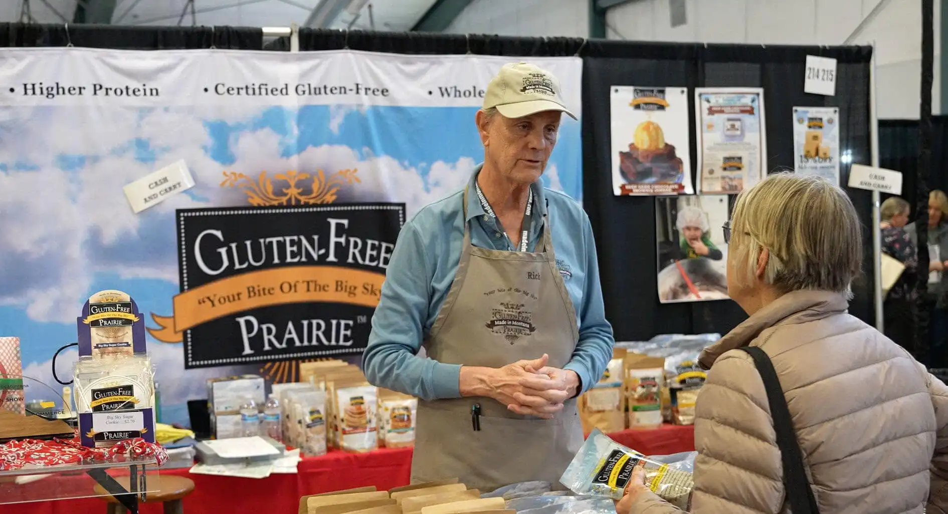 A vendor in a beige apron and cap talks to a customer at a booth labeled Gluten-Free Prairie during the Made in Montana Tradeshow. Various gluten-free products from Montana businesses are displayed, with banners and packages visible in the background.