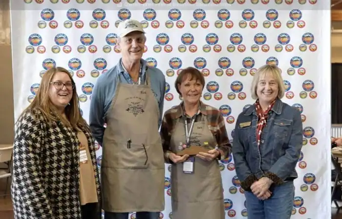 Four smiling adults stand indoors in front of a white step-and-repeat banner with colorful round logos. At center, two people from Gluten-Free Prairie wear aprons and hold trophies, celebrating quality and community. Tables and chairs are visible in the background.