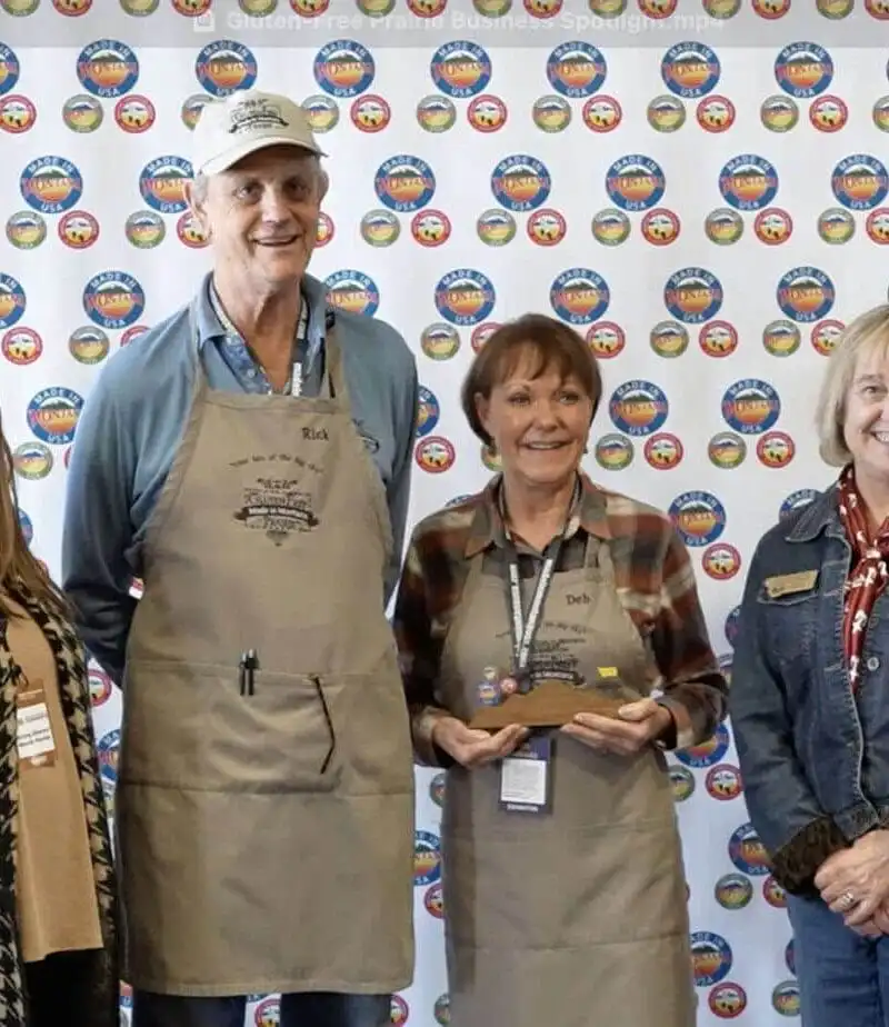 Four smiling adults stand indoors in front of a white step-and-repeat banner with colorful round logos. At center, two people from Gluten-Free Prairie wear aprons and hold trophies, celebrating quality and community. Tables and chairs are visible in the background.