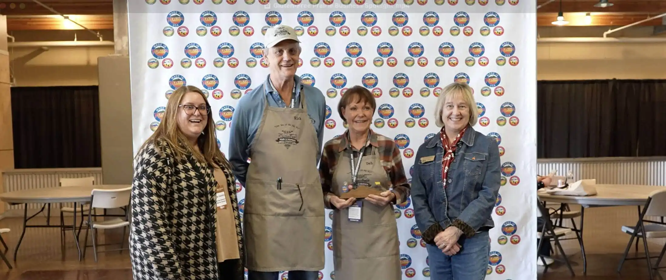 Four smiling adults stand indoors in front of a white step-and-repeat banner with colorful round logos. At center, two people from Gluten-Free Prairie wear aprons and hold trophies, celebrating quality and community. Tables and chairs are visible in the background.