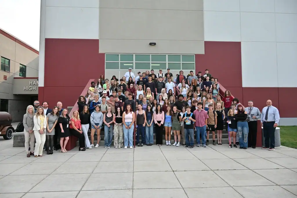 A large group of students and adults, including Secretary Christi Jacobsen, pose together on steps outside a school building, smiling for a group photo.