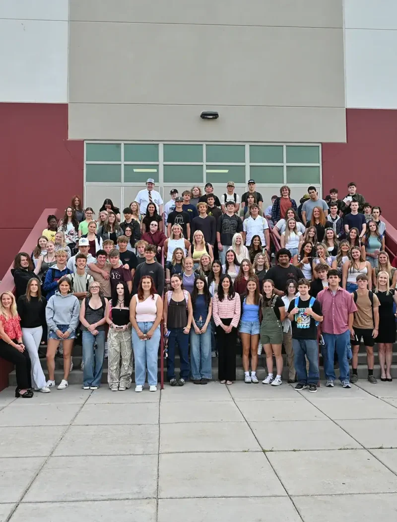 A large group of students and adults, including Secretary Christi Jacobsen, pose together on steps outside a school building, smiling for a group photo.