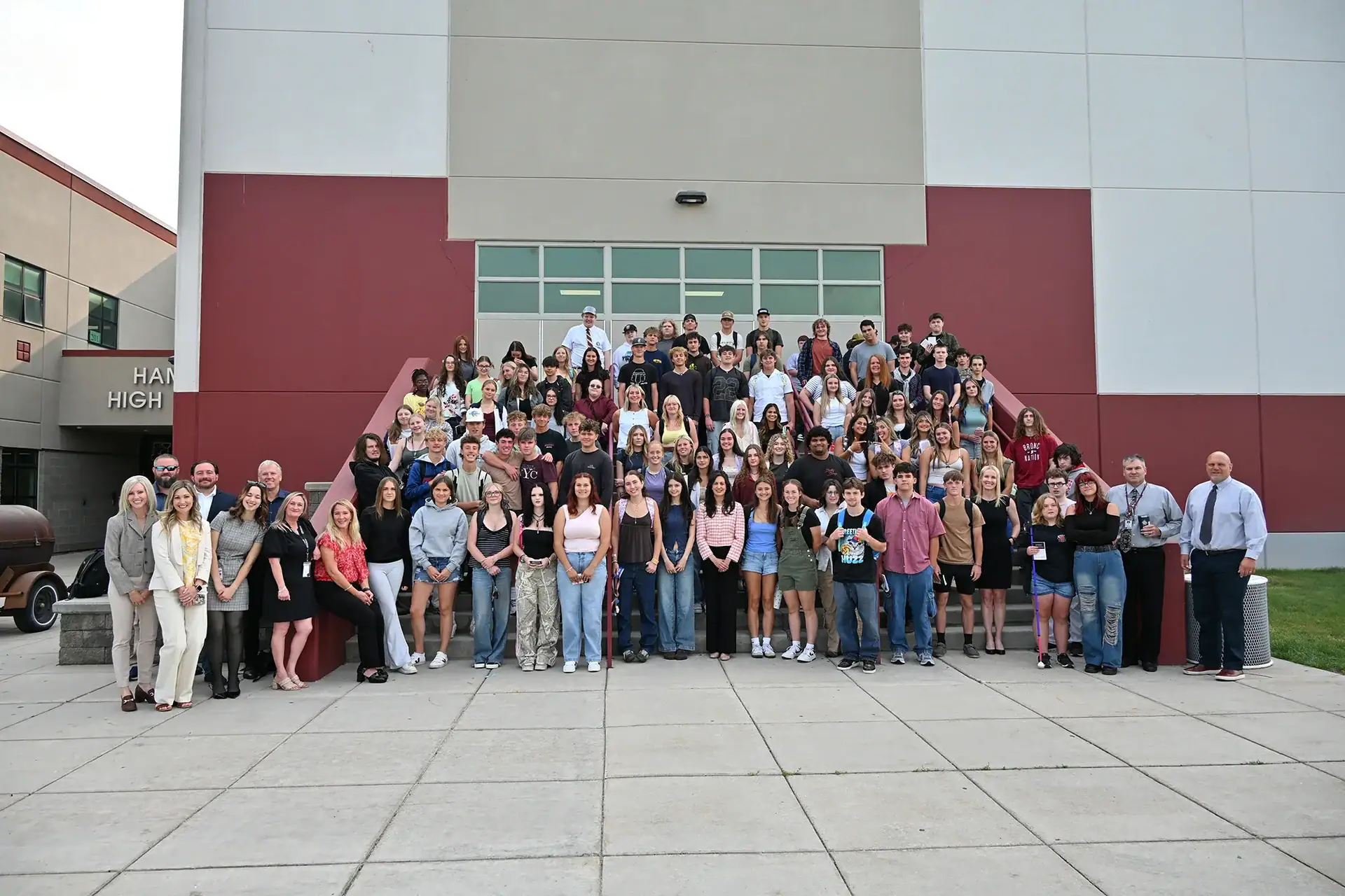 A large group of students and adults, including Secretary Christi Jacobsen, pose together on steps outside a school building, smiling for a group photo.