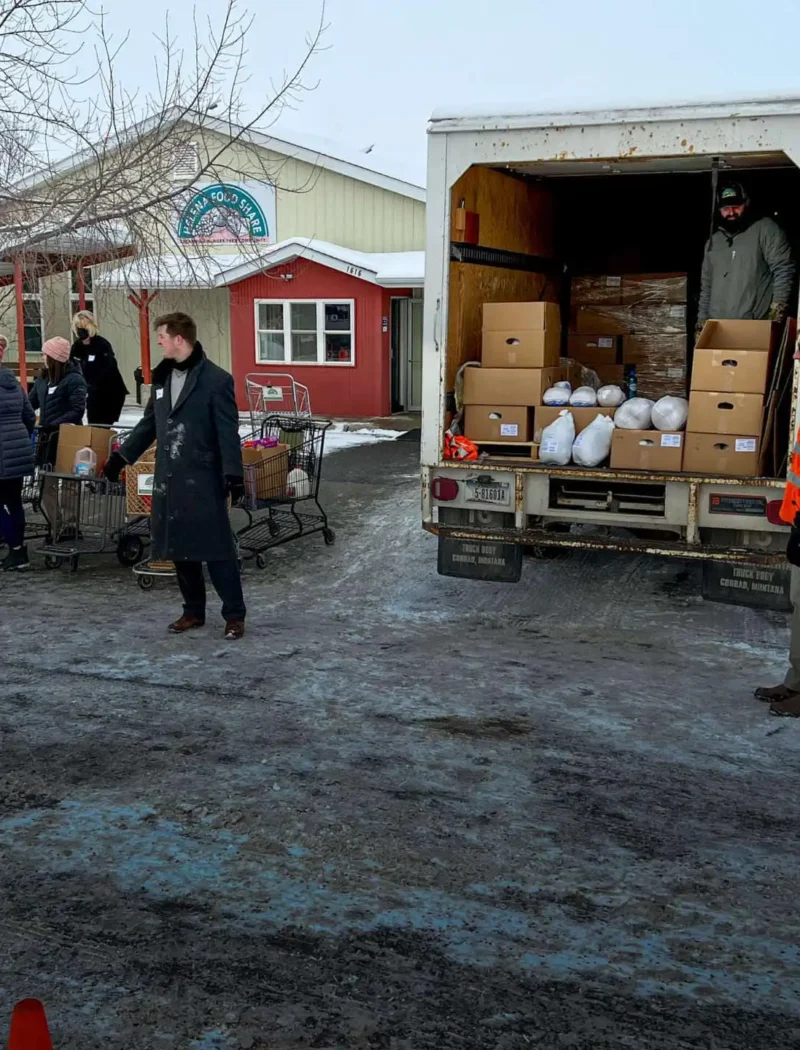 People in winter clothing stand near a truck unloading boxes and bags for Helena Food Share in a snowy parking lot, with some loading holiday meals onto carts. Orange cones and two workers in safety vests are directing the activity.