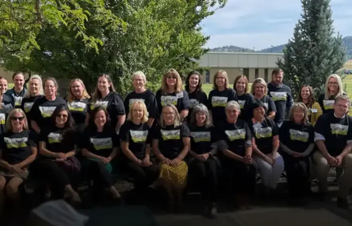 Secretary of State's Elections Division concludes Helena-area training 9 A group of about 30 adults, mostly women, from an Elections Division Helena-area training pose outdoors together in matching black shirts with a yellow and white graphic. They are sitting and standing on a patio with trees and grass in the background.