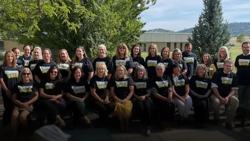 A group of about 30 adults, mostly women, from an Elections Division Helena-area training pose outdoors together in matching black shirts with a yellow and white graphic. They are sitting and standing on a patio with trees and grass in the background.
