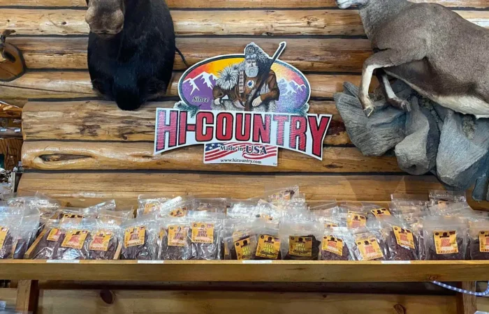 Montana Business Spotlight: Inspiring innovations at Hi Country Snack Foods 8 Packets of jerky are lined up on a wooden shelf with a “Hi Country” sign above, featuring a frontiersman and mountains. Taxidermy animals are mounted on the log cabin-style wall.