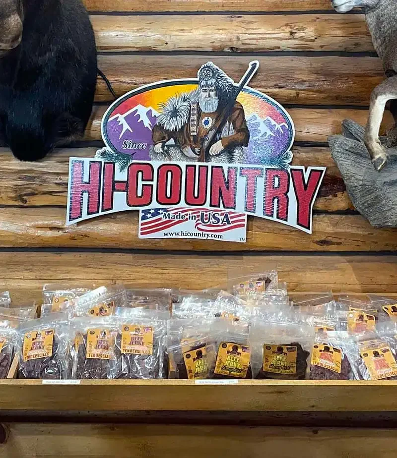 Packets of jerky are lined up on a wooden shelf with a “Hi Country” sign above, featuring a frontiersman and mountains. Taxidermy animals are mounted on the log cabin-style wall.