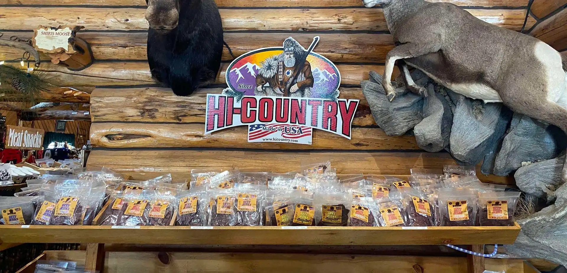 Packets of jerky are lined up on a wooden shelf with a “Hi Country” sign above, featuring a frontiersman and mountains. Taxidermy animals are mounted on the log cabin-style wall.