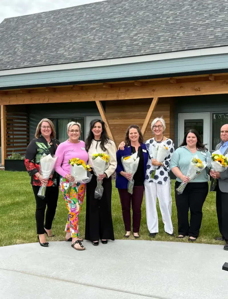 Seven people stand in a row outside a modern Montana building, each holding a bouquet of yellow flowers and smiling at the camera. The group is dressed in business casual attire, perhaps celebrating recent business registrations.