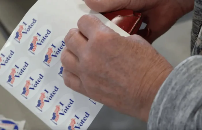 Montana Secretary of State Christi Jacobsen celebrates successful 2022 Primary Election 7 A person in a gray long-sleeve shirt peels off an I Voted sticker with an American flag, celebrating participation in the 2022 Primary Election overseen by Montana Secretary of State Christi Jacobsen.