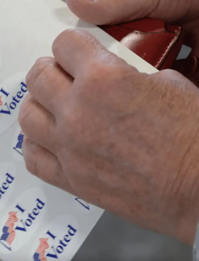 A person in a gray long-sleeve shirt peels off an I Voted sticker with an American flag, celebrating participation in the 2022 Primary Election overseen by Montana Secretary of State Christi Jacobsen.
