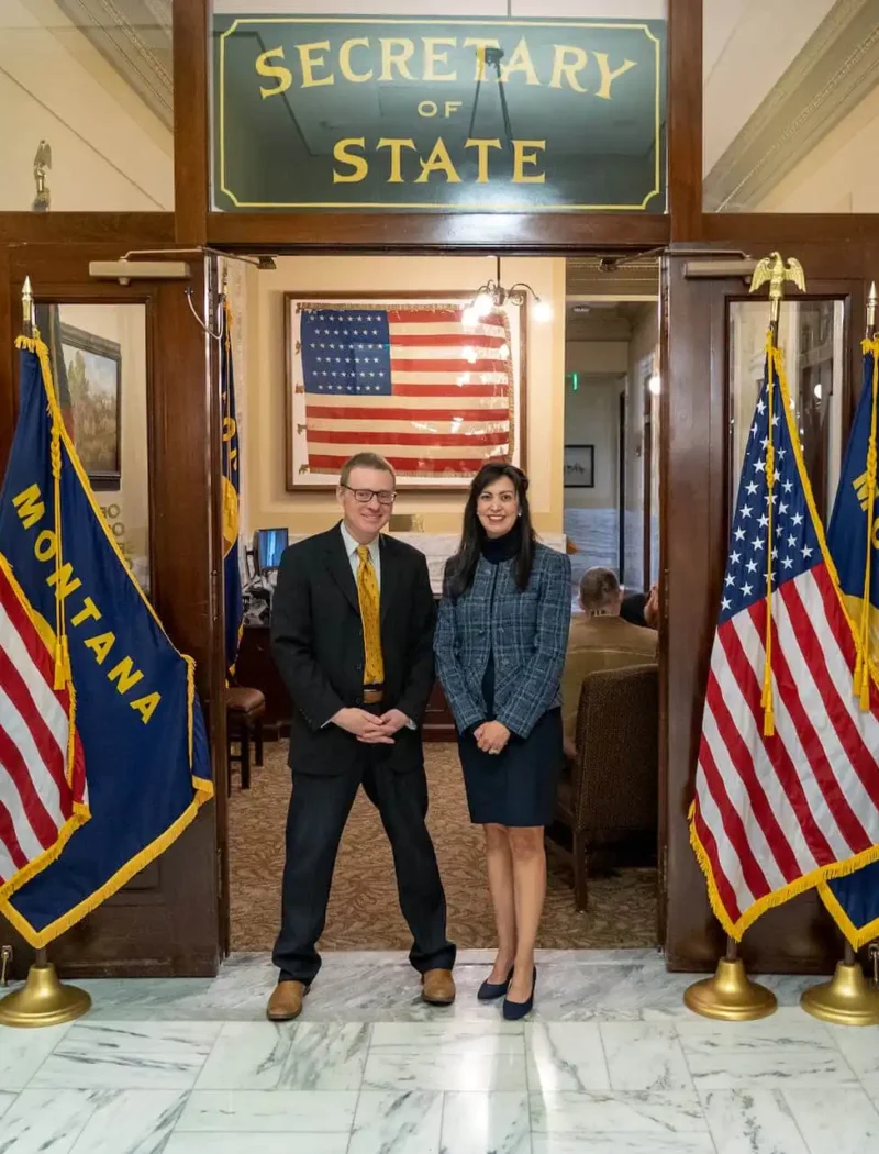 Two people stand in front of Montana and U.S. flags at the entrance to the Secretary of State office. An American flag is displayed inside, and the area has marble floors and wood-paneled doors.
