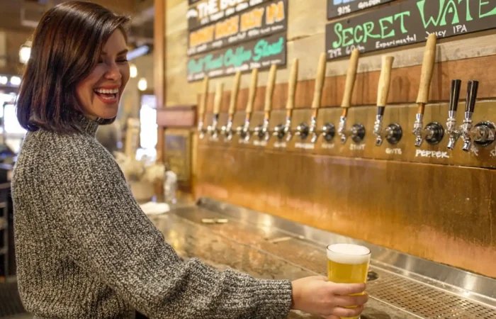 Montana Business Spotlight: Sacred Waters Brewing Co. sharing Montana through craft 1 A woman with short dark hair smiles while holding a pint of Montana craft beer in front of several wooden tap handles at a bar, with chalkboard menus in the background.