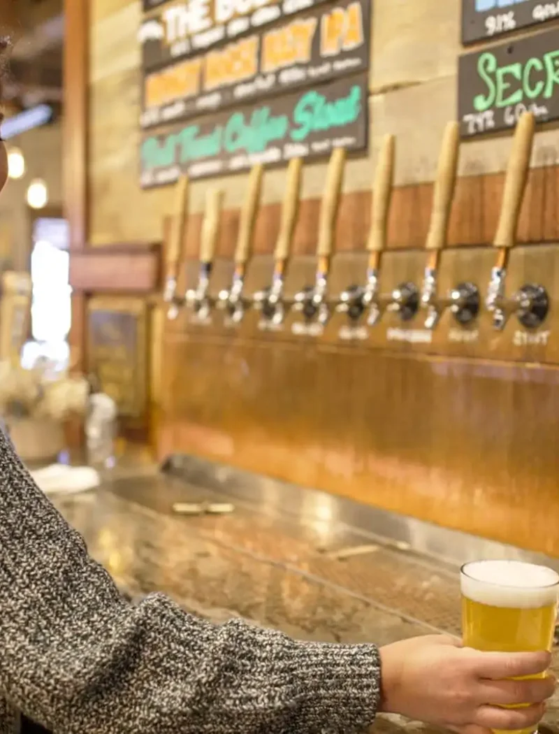 A woman with short dark hair smiles while holding a pint of Montana craft beer in front of several wooden tap handles at a bar, with chalkboard menus in the background.