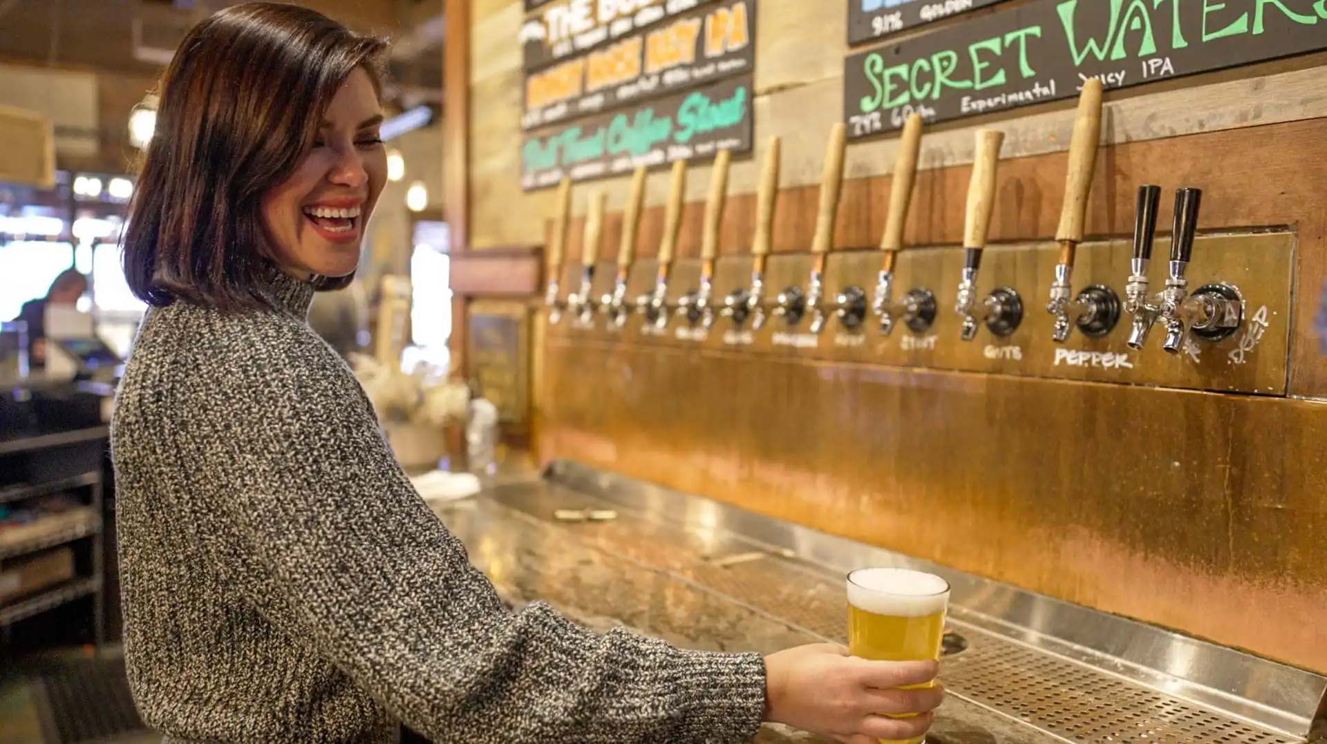 A woman with short dark hair smiles while holding a pint of Montana craft beer in front of several wooden tap handles at a bar, with chalkboard menus in the background.