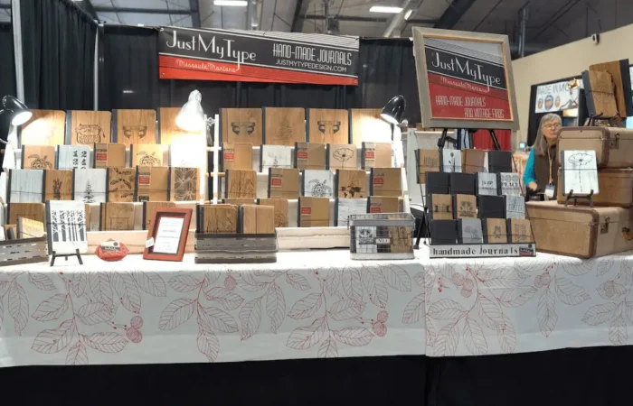 Montana Business Spotlight: Creativity encouraged in Just My Type handmade journals and sketchbooks 4 A market stall from a Montana business displays handmade journals and sketchbooks with various designs on a table covered by a white cloth with red leaf patterns. The booth sign reads Just My Type Handmade Journals. Suitcases and lamps are also visible.