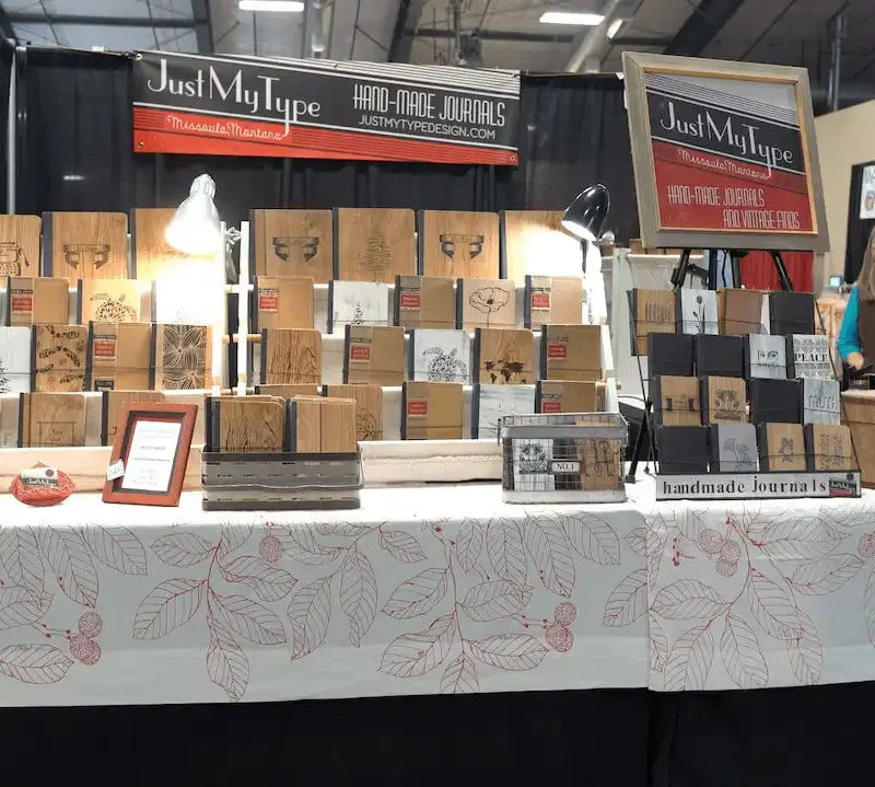 A market stall from a Montana business displays handmade journals and sketchbooks with various designs on a table covered by a white cloth with red leaf patterns. The booth sign reads Just My Type Handmade Journals. Suitcases and lamps are also visible.