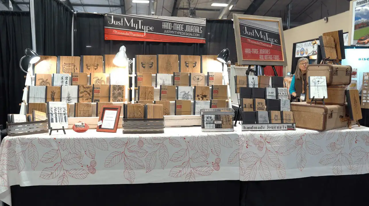 A market stall from a Montana business displays handmade journals and sketchbooks with various designs on a table covered by a white cloth with red leaf patterns. The booth sign reads Just My Type Handmade Journals. Suitcases and lamps are also visible.