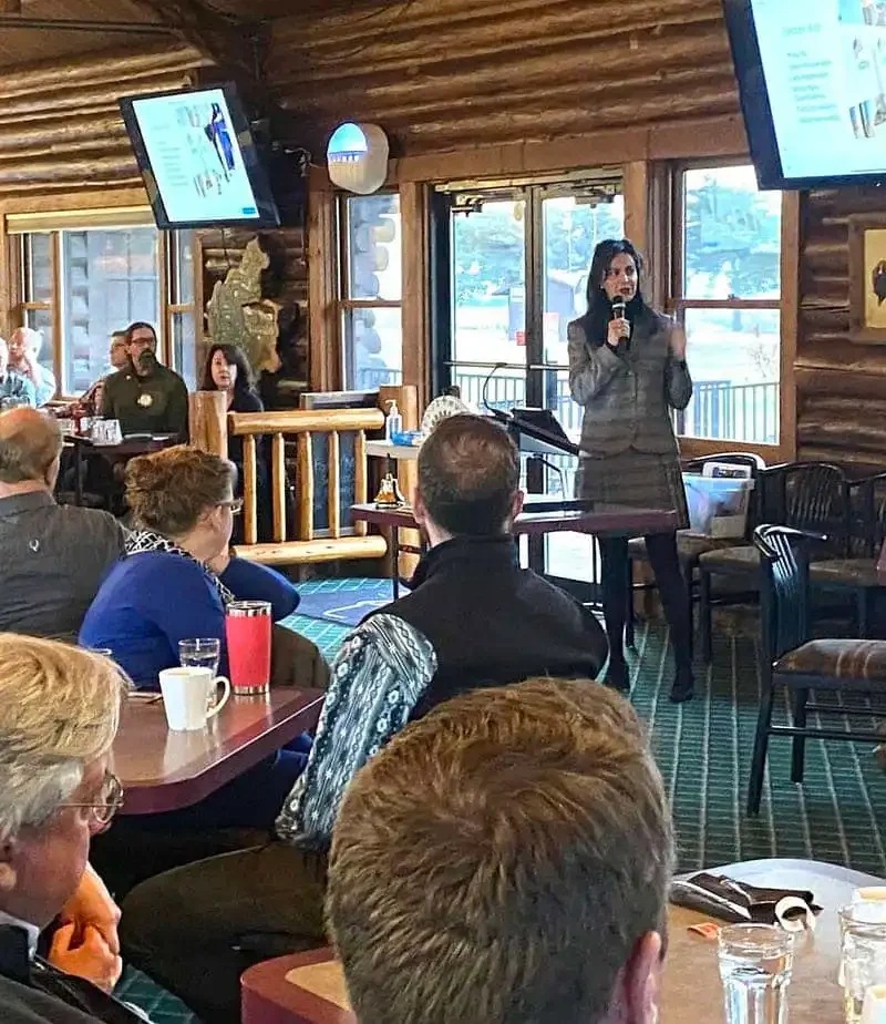 A woman speaks with a microphone in front of a group of people seated at tables in a rustic, log cabin-style room. A presentation on new business registrations is displayed on screens behind her, featuring Christi Jacobsen’s March report.