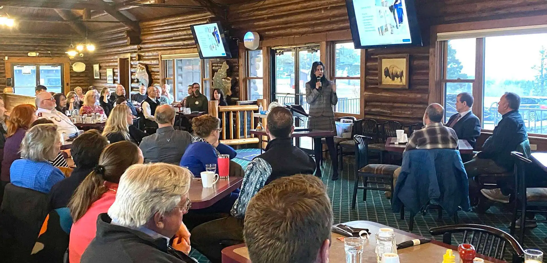 A woman speaks with a microphone in front of a group of people seated at tables in a rustic, log cabin-style room. A presentation on new business registrations is displayed on screens behind her, featuring Christi Jacobsen’s March report.