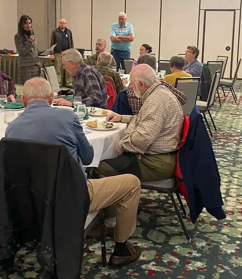 A group of people sit around tables in a conference room, listening to Christi Jacobsen speak at the front. Some are eating while others watch attentively, discussing annual report filing for Montana businesses. The room has patterned carpet and neutral walls.