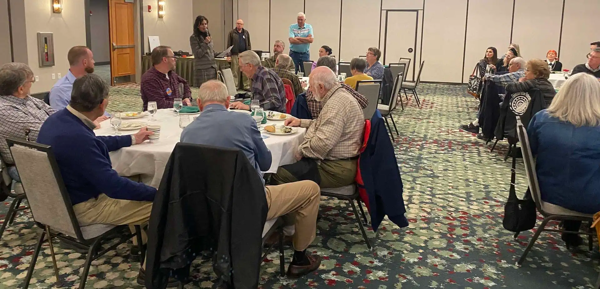 A group of people sit around tables in a conference room, listening to Christi Jacobsen speak at the front. Some are eating while others watch attentively, discussing annual report filing for Montana businesses. The room has patterned carpet and neutral walls.