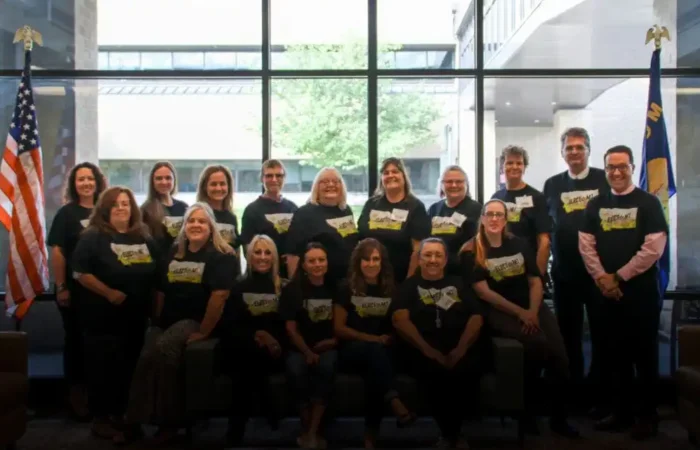 Elections Division wraps up electMT training in Kalispell 8 A group of sixteen adults pose indoors in front of large windows, wearing matching dark t-shirts with yellow designs during electMT training. Two flags are displayed at the edges, and trees are visible outside in Kalispell.