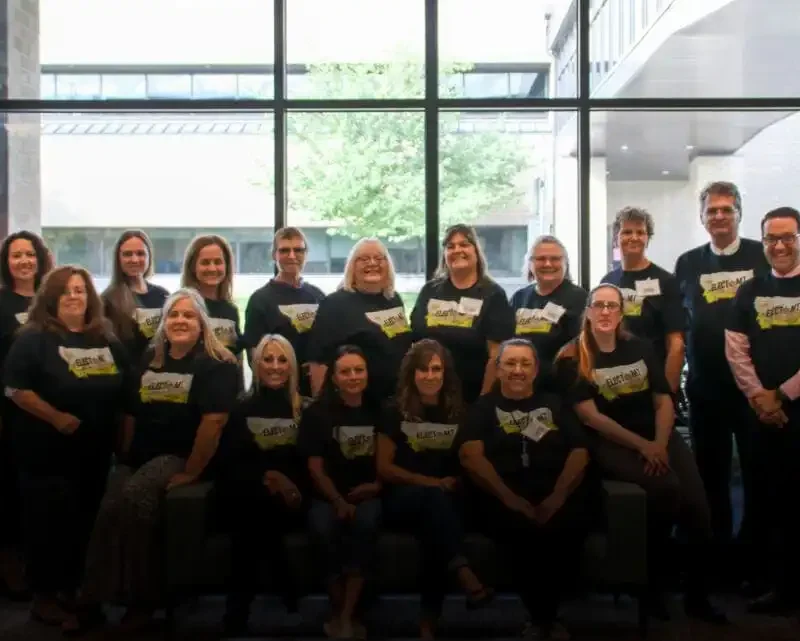Elections Division wraps up electMT training in Kalispell 9 A group of sixteen adults pose indoors in front of large windows, wearing matching dark t-shirts with yellow designs during electMT training. Two flags are displayed at the edges, and trees are visible outside in Kalispell.