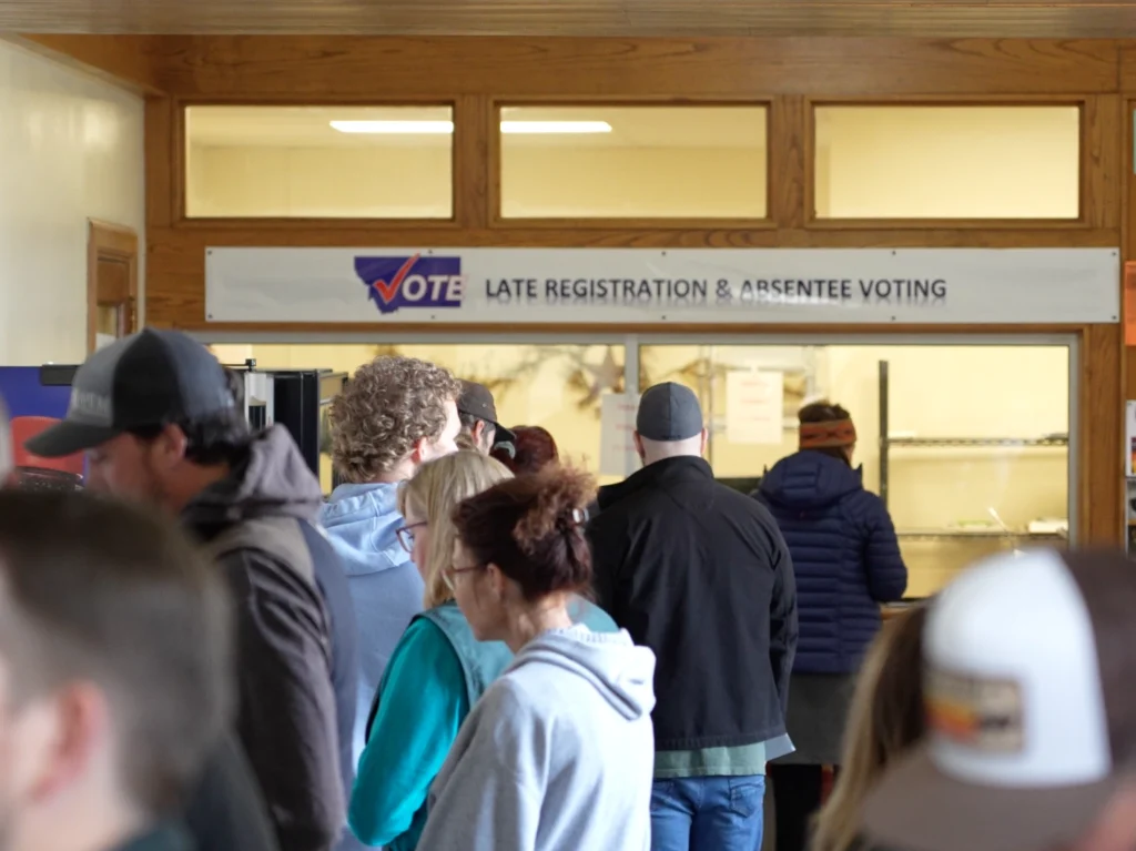 People stand in line at an indoor polling place near a sign that reads Vote - Late Registration & Absentee Voting, reflecting the busy atmosphere of the Montana Primary Election at local county election offices.