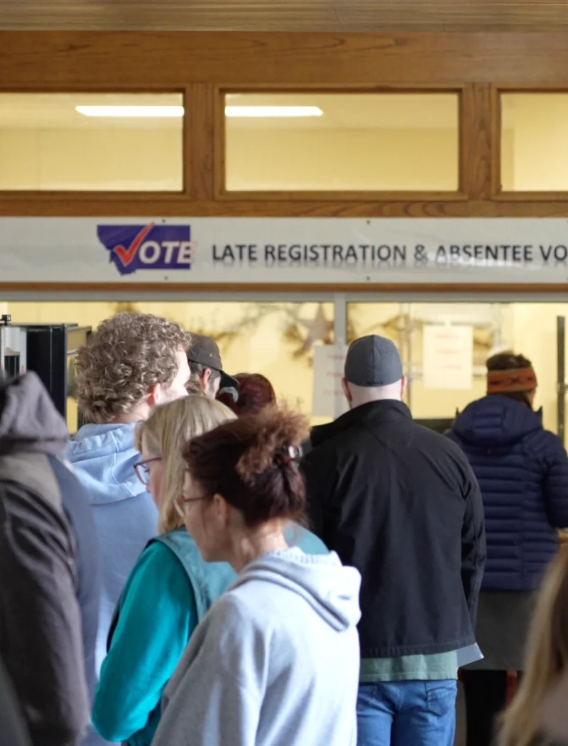 People stand in line at an indoor polling place near a sign that reads Vote - Late Registration & Absentee Voting, reflecting the busy atmosphere of the Montana Primary Election at local county election offices.
