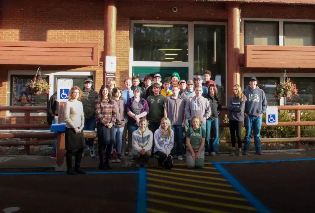 A large group of people pose for a photo outside Lincoln High School, a brick building with large windows. Some are standing, others kneeling in front, near accessible parking signs and hanging flower baskets. Secretary Jacobsen is also present.