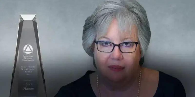 A woman with gray hair and glasses sits next to a tall, clear achievement award trophy against a gray background. The text on the trophy references Lori Hamm and the National Notary Association.