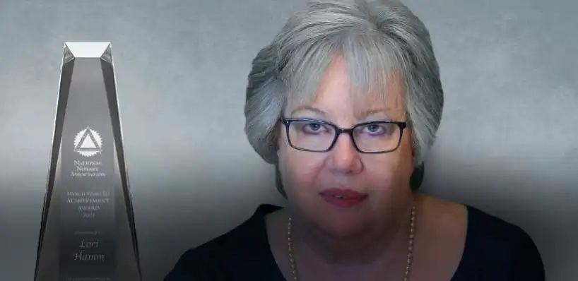 A woman with gray hair and glasses sits next to a tall, clear achievement award trophy against a gray background. The text on the trophy references Lori Hamm and the National Notary Association.