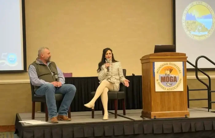 Secretary Jacobsen highlights red tape reduction with Montana Outfitters & Guides Association 2 A man and a woman are seated on a stage, with the woman speaking into a microphone. An empty podium with the Montana Outfitters and Guides Association (MOGA) logo and a large seal are visible in the background, highlighting efforts in red tape reduction.
