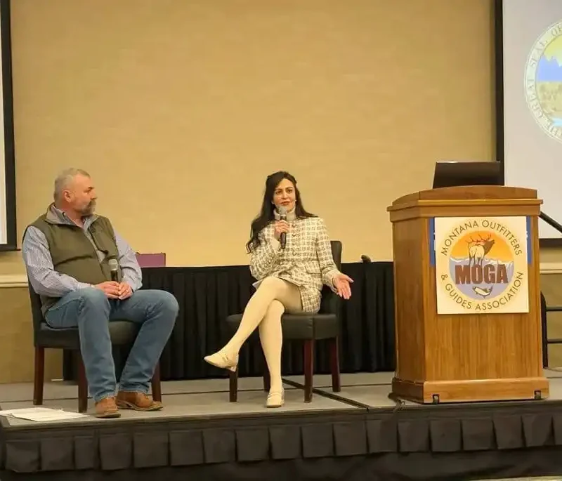 A man and a woman are seated on a stage, with the woman speaking into a microphone. An empty podium with the Montana Outfitters and Guides Association (MOGA) logo and a large seal are visible in the background, highlighting efforts in red tape reduction.
