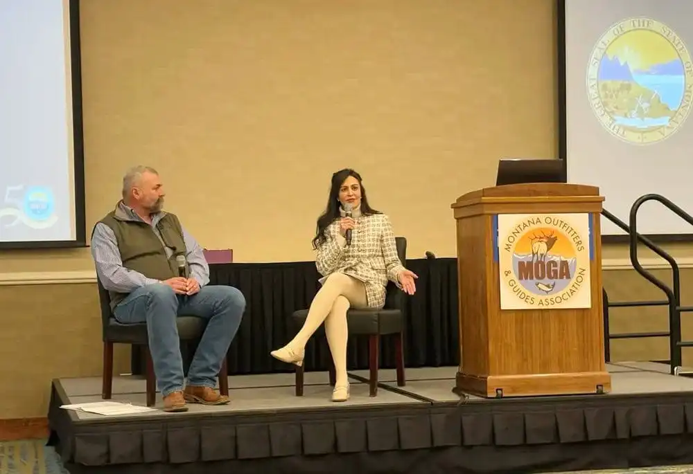A man and a woman are seated on a stage, with the woman speaking into a microphone. An empty podium with the Montana Outfitters and Guides Association (MOGA) logo and a large seal are visible in the background, highlighting efforts in red tape reduction.