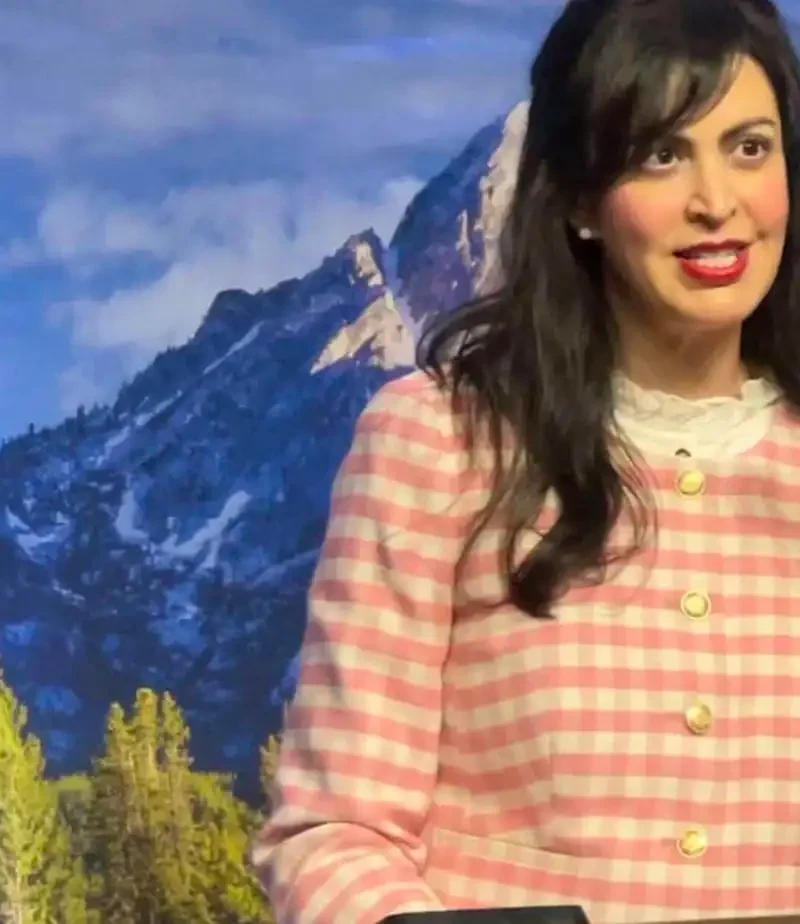 Christi Jacobsen, Montana Secretary of State, stands in a pink and white checkered jacket at a podium with a microphone, addressing new business registrations against a scenic backdrop of mountains and pine trees.