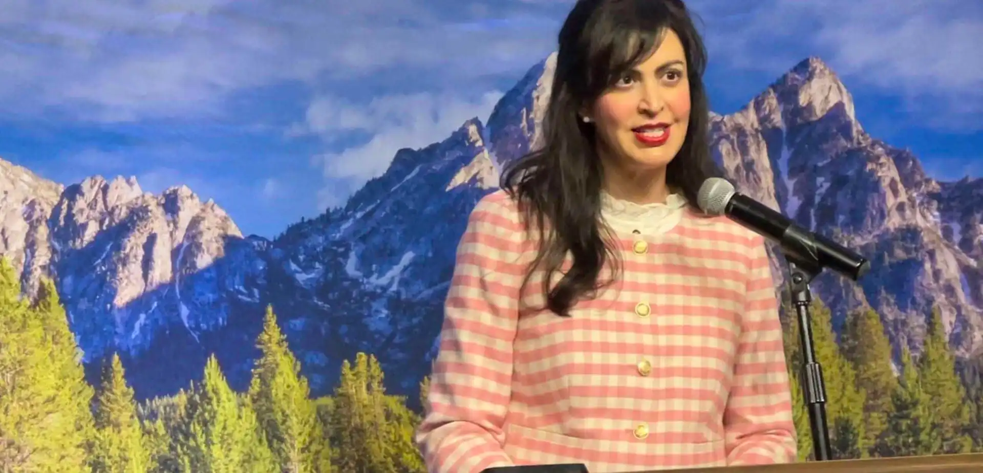 Christi Jacobsen, Montana Secretary of State, stands in a pink and white checkered jacket at a podium with a microphone, addressing new business registrations against a scenic backdrop of mountains and pine trees.